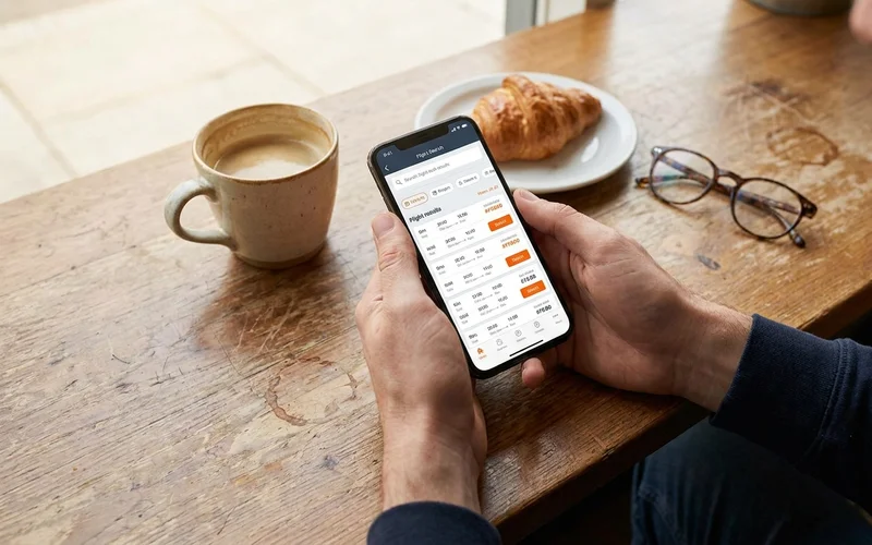 A realistic overhead shot of a person's hands holding a smartphone showing a flight search results page at a coffee shop table, warm natural lighting, 16:9 composition. Alt: person searching for cheapest days to fly on a smartphone.